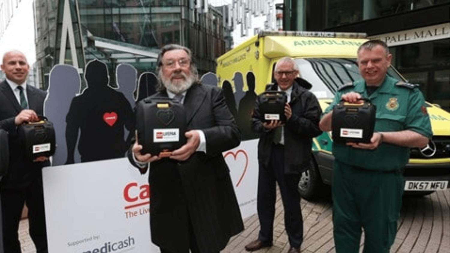 Four males stood in front of an ambulance holding a defibrillator