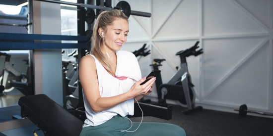woman sat on weights bench in the gym holding smartphone with headphones in and smiling