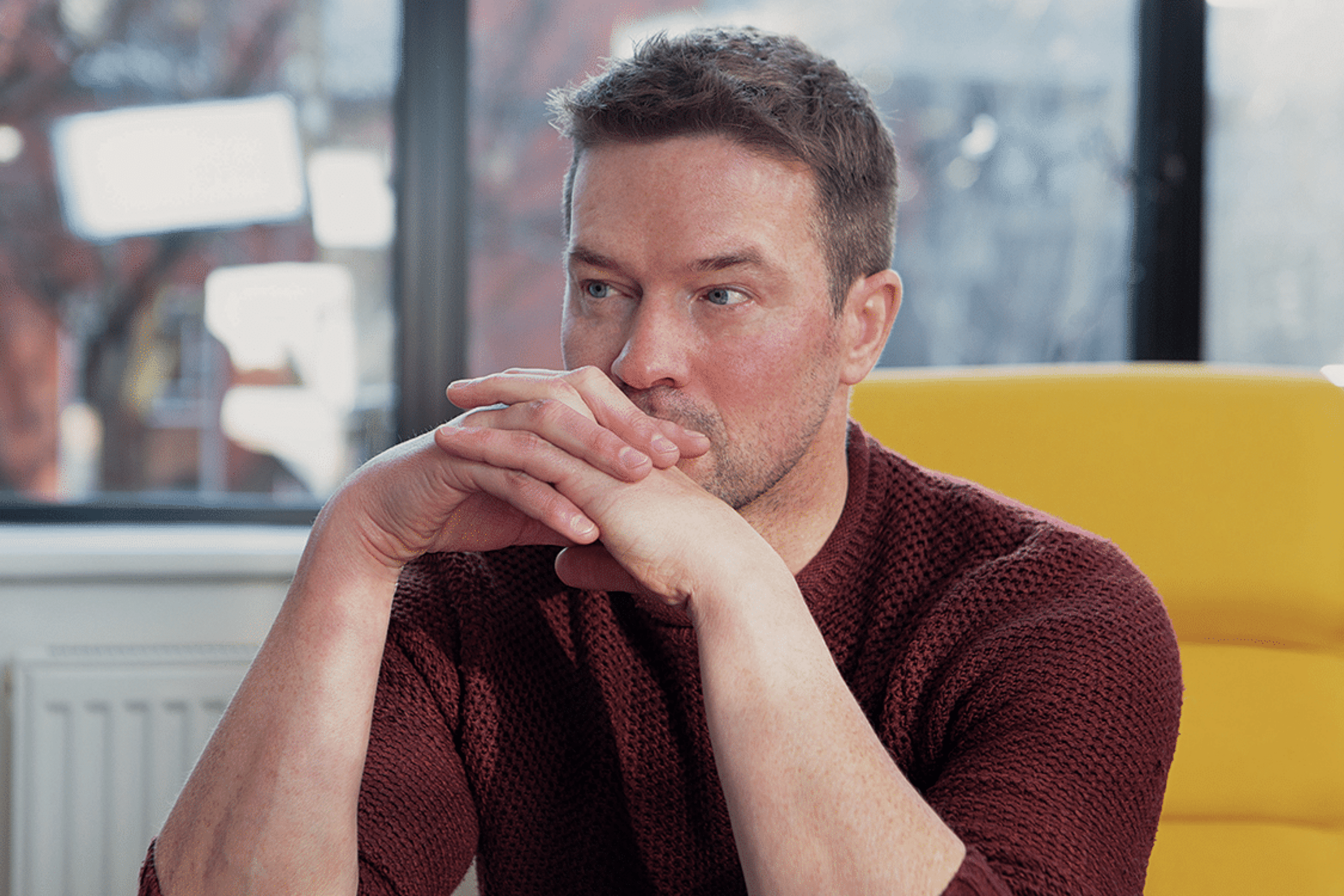 man in burgundy jumper resting is chin on clasped hands deep in thought