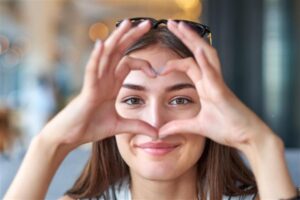 Woman making a heart with her hands so that her eyes show but the rest of her face is covered.