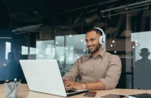 male employee sat in an office environment looking and smiling at laptop with headphones on